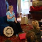 Photo of Betty sitting in her storytelling chair with a drum at her feet and a captive audience